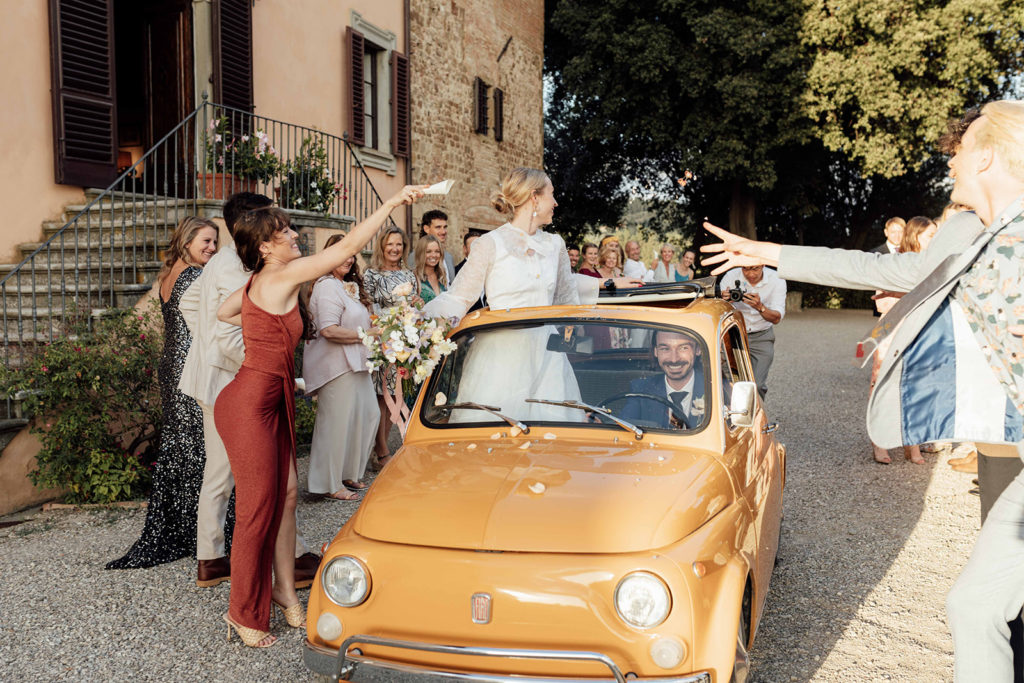 Bride and groom driving a vintage Fiat 500 are showered by confetti by their wedding guests 