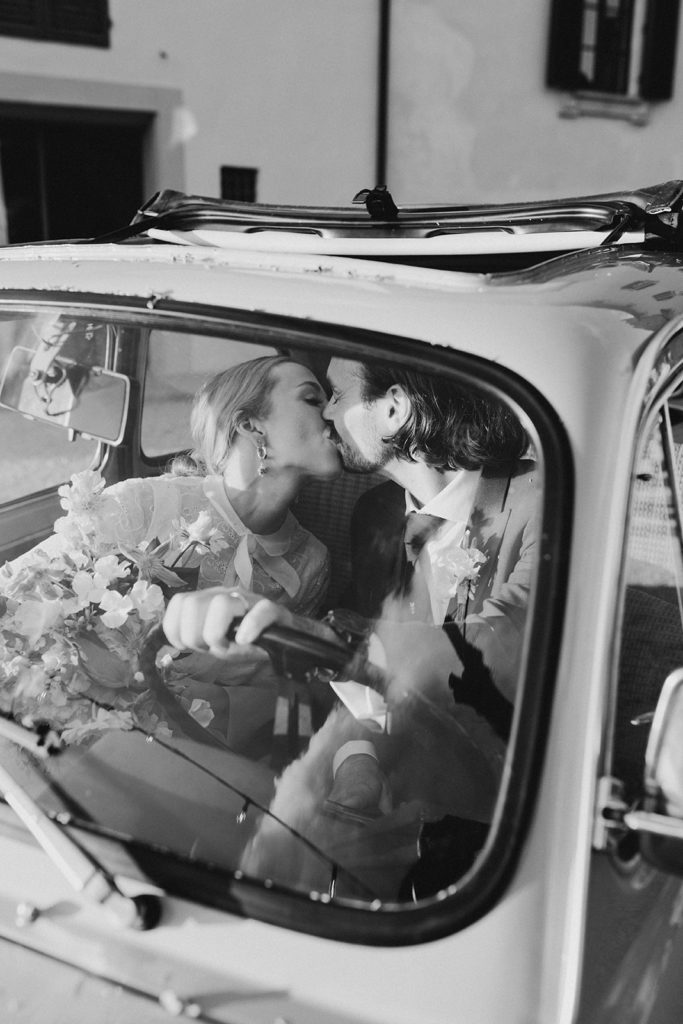 Bride and groom share a kiss at the while of a vintage Fiat 500 car.
