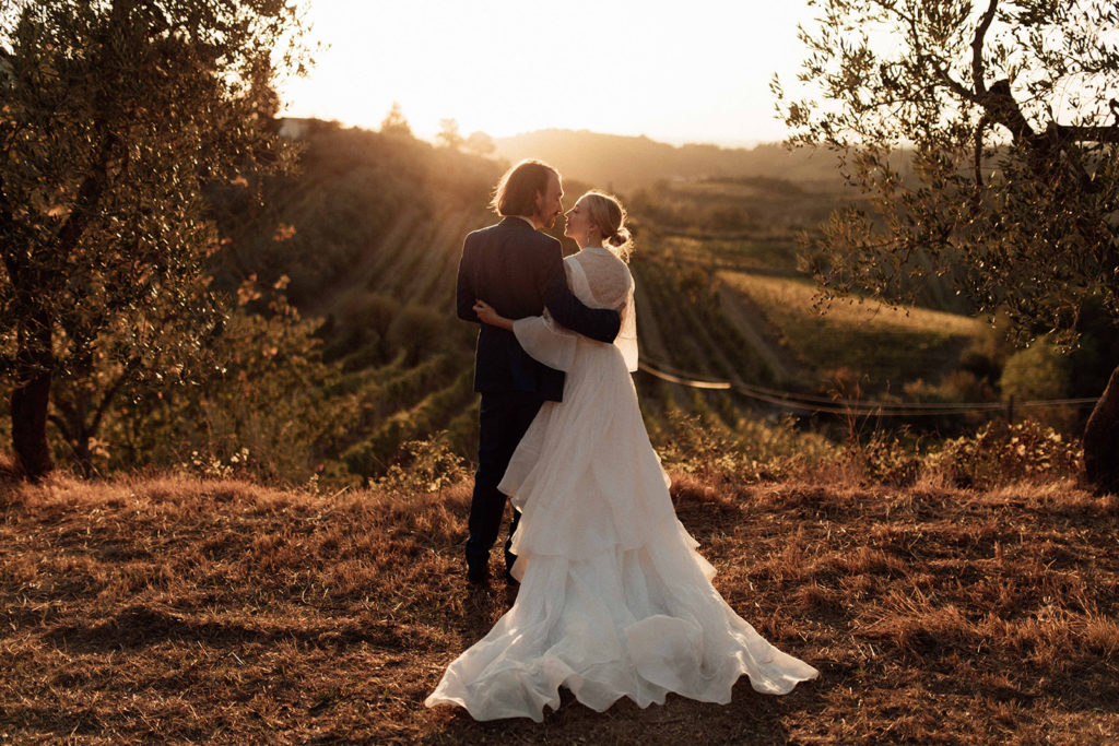 bride and groom look out over a golden Tuscan sunset in italy. the bride wears a long white gown and the groom wears a blue suit.