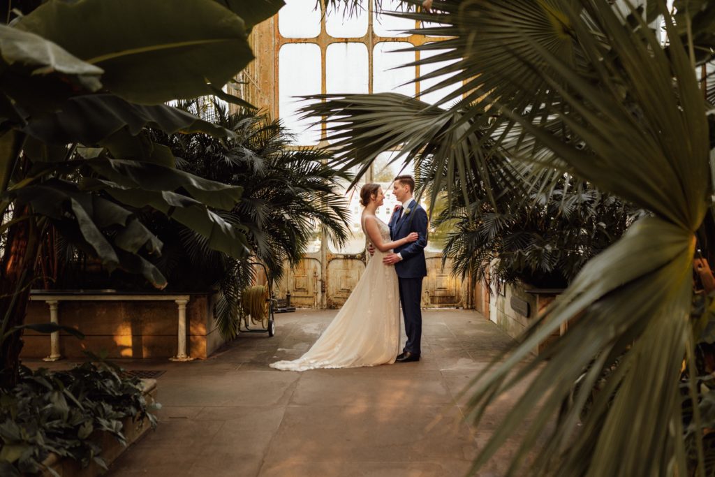 A bride and groom stand facing each other in a palm house conservatory on their wedding day. 